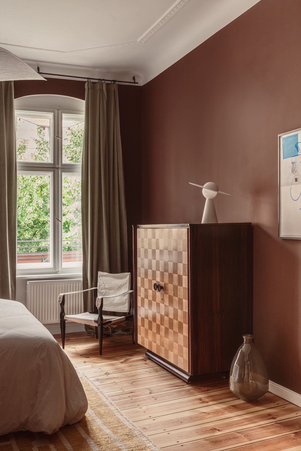 The bedroom with oxblood walls, an arched window with trees outside, and a tall checkerboard-front wardrobe.