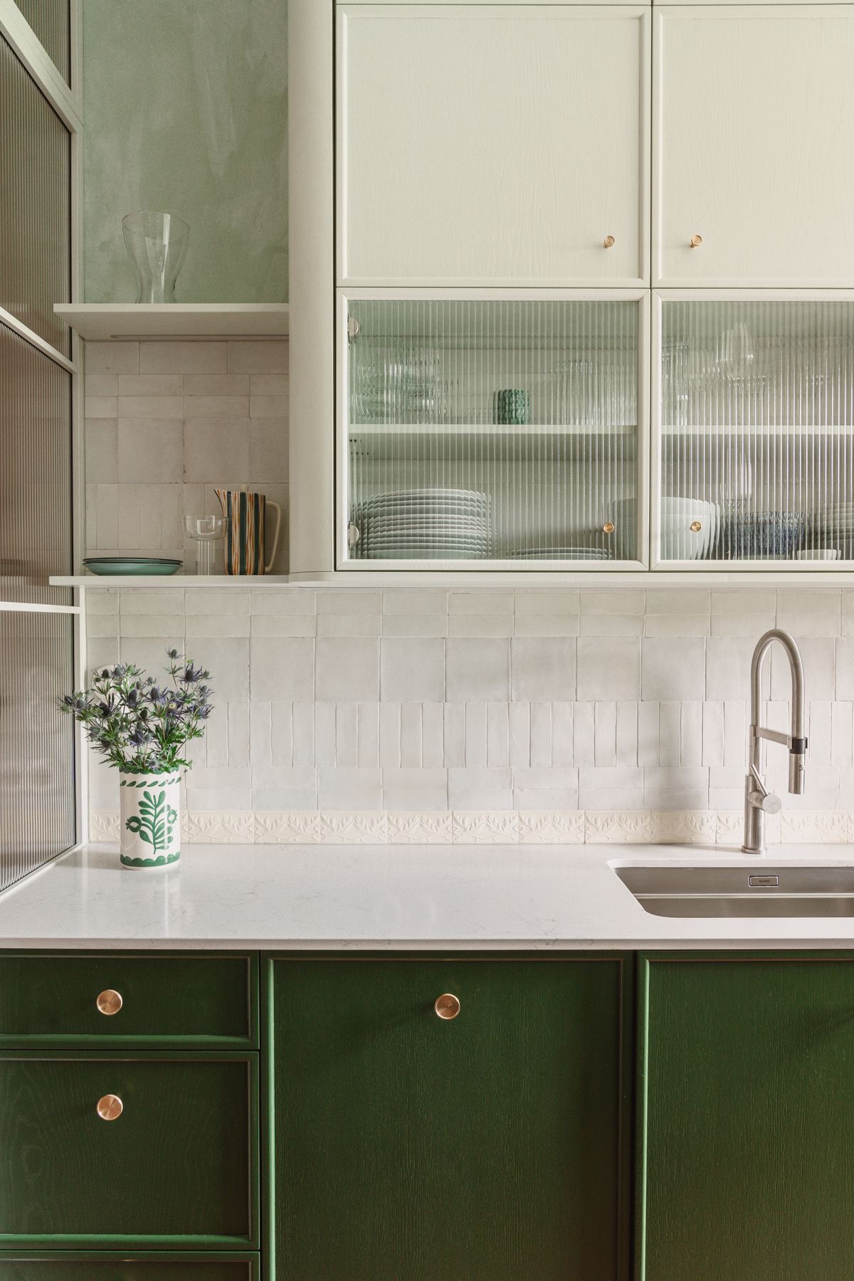 The kitchen counter with green cabinets below, white tiles above it, and glass-fronted cupboards holding stacked plates.
